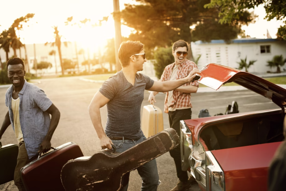 Three young men packing a car with suitcases and a guitar for a road trip